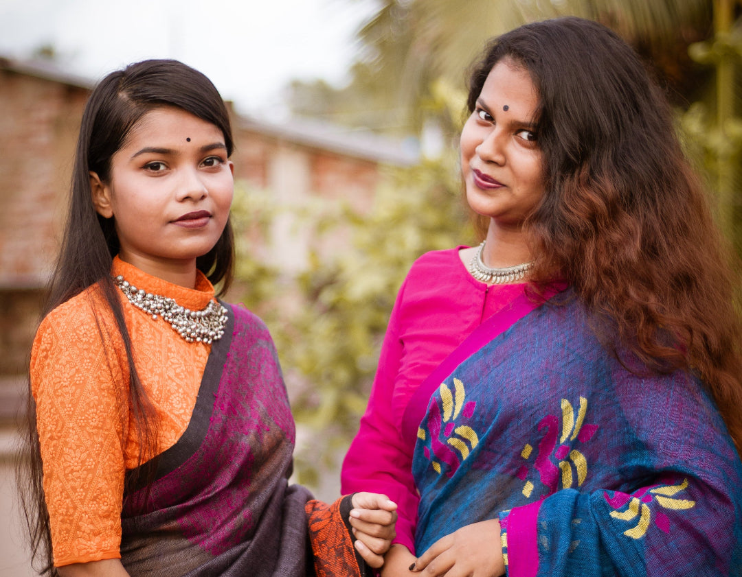 Two women standing outdoors wearing traditional Indian sarees. The woman on the left wears an orange and dark purple saree, while the woman on the right wears a bright pink blouse and a blue saree with yellow and purple patterns.